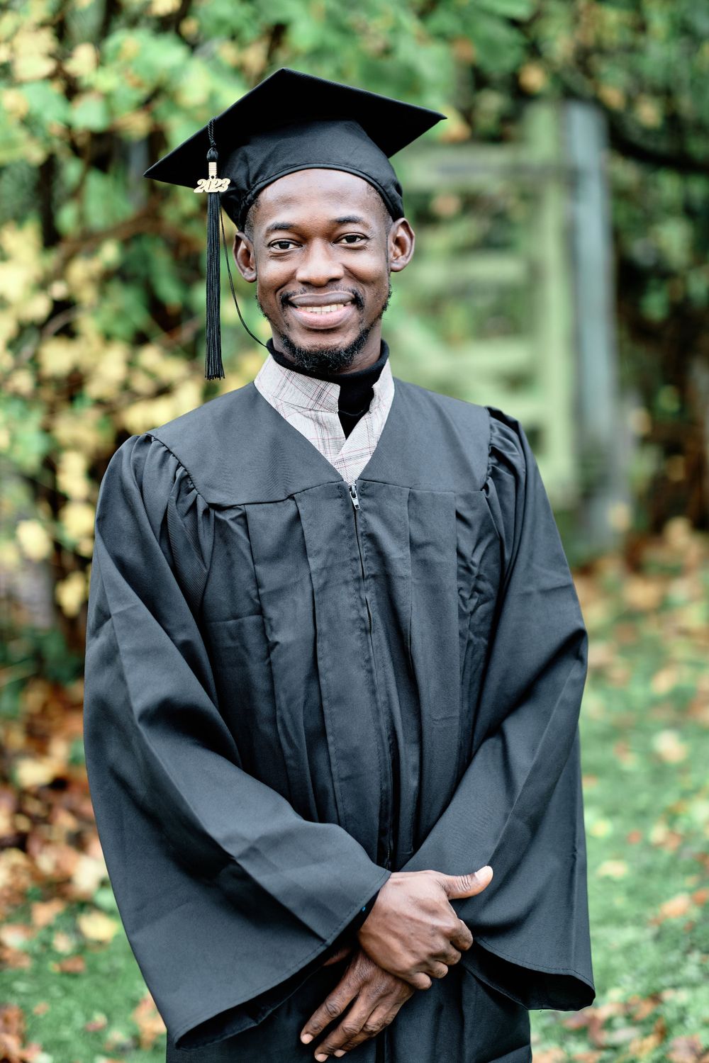 Graduate in traditional black commencement regalia smiling against leafy background.