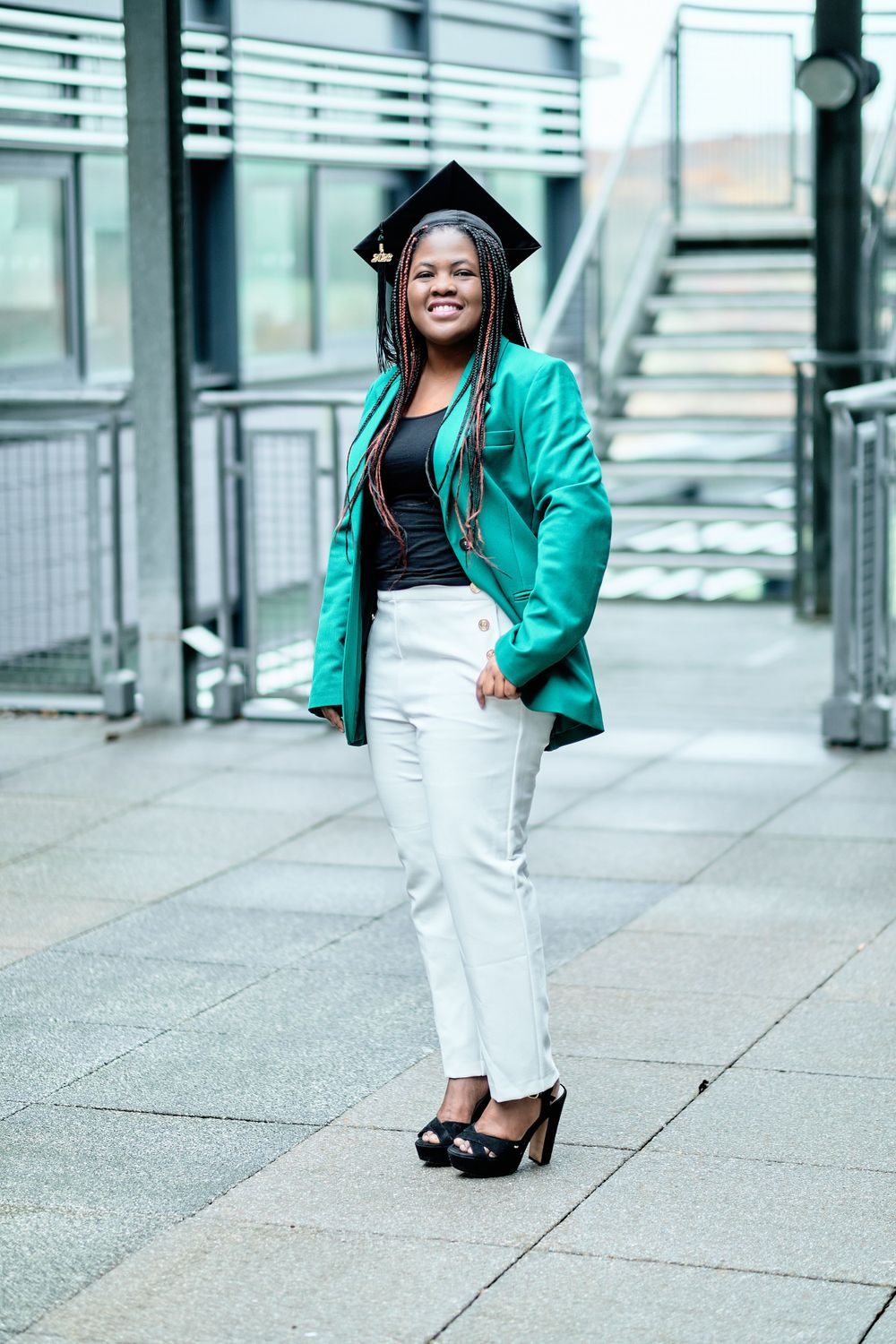 Graduate wearing teal blazer and graduation cap poses confidently on modern architecture walkway.