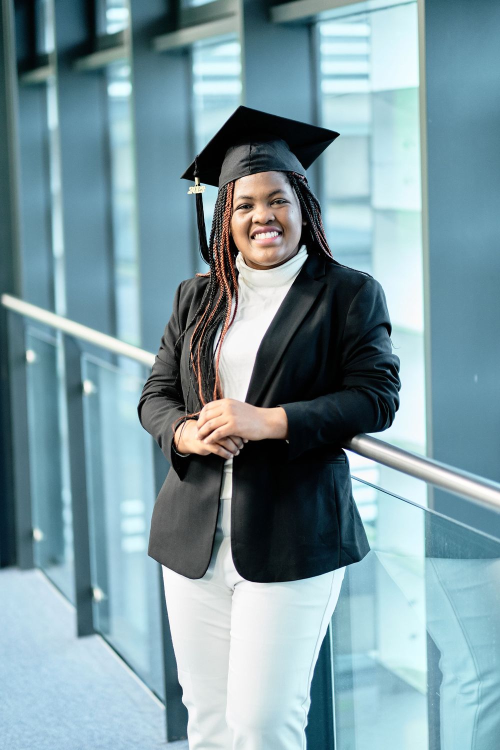 Graduate wearing graduation cap poses in modern building with white turtleneck and black blazer.