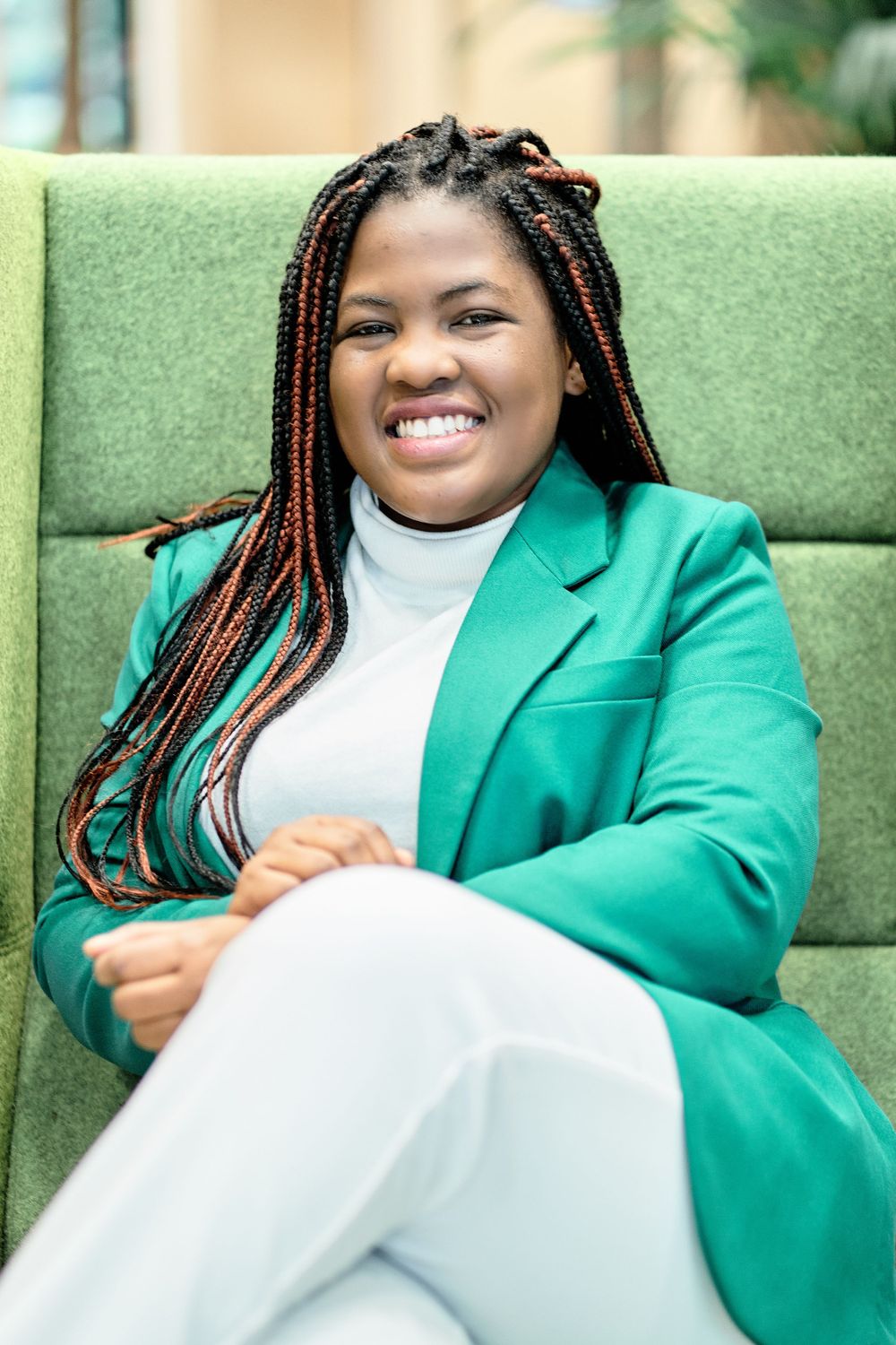 Professional in emerald blazer relaxes on green couch wearing white outfit.