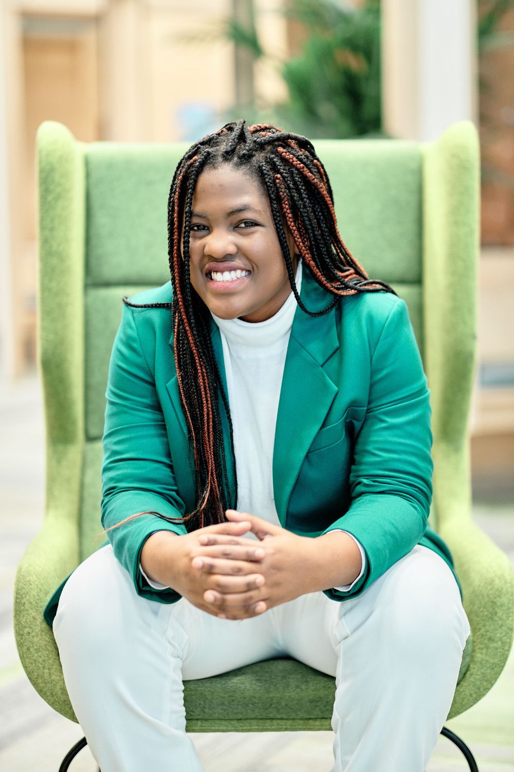 Professional in green blazer sits casually in wingback chair with bright smile.