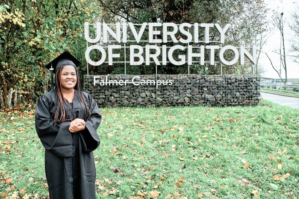 Graduate poses in front of University of Brighton campus sign during graduation celebration.