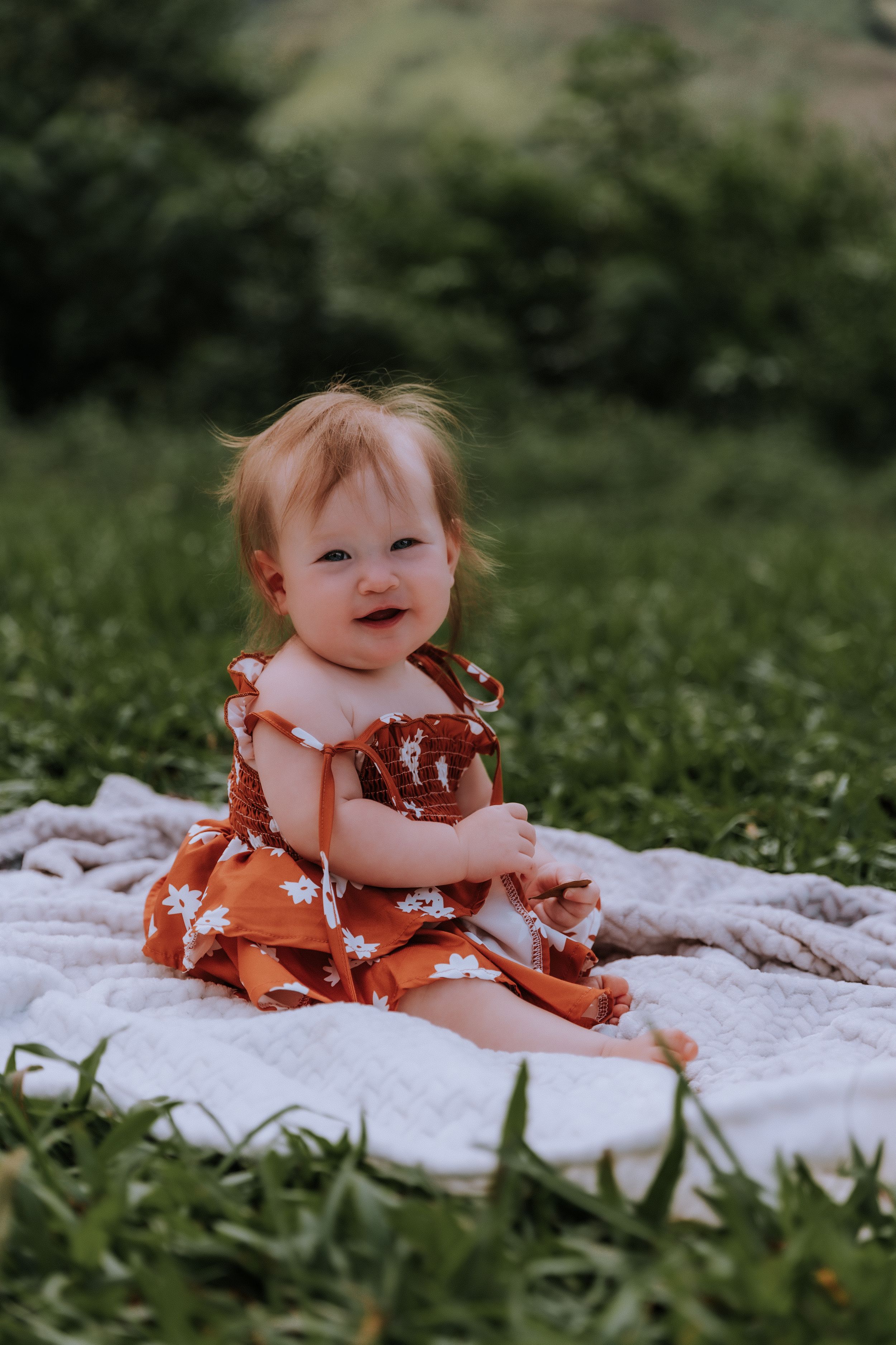 Baby in a rust-colored dress sits smiling on a white blanket in a grassy field.