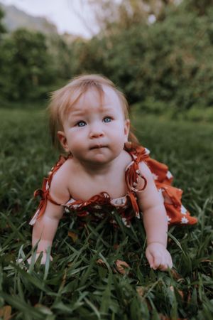 A baby in a red floral dress sits in lush grass with majestic mountains in the background.