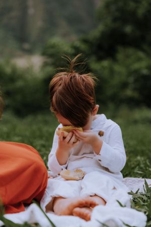 Child eating snack while sitting on white blanket during outdoor picnic.