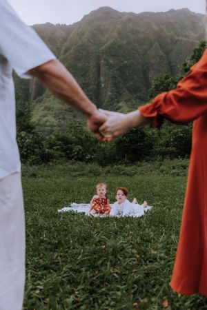 A couple holds hands while watching children on a blanket against a backdrop of lush mountain scenery.