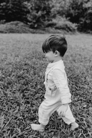 Black and white portrait of toddler walking in field wearing white outfit.