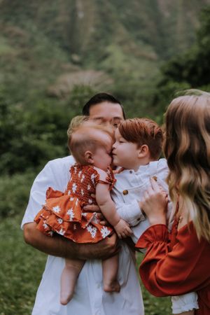 A family shares tender kisses in coordinated white and rust-colored outfits against a natural outdoor backdrop.