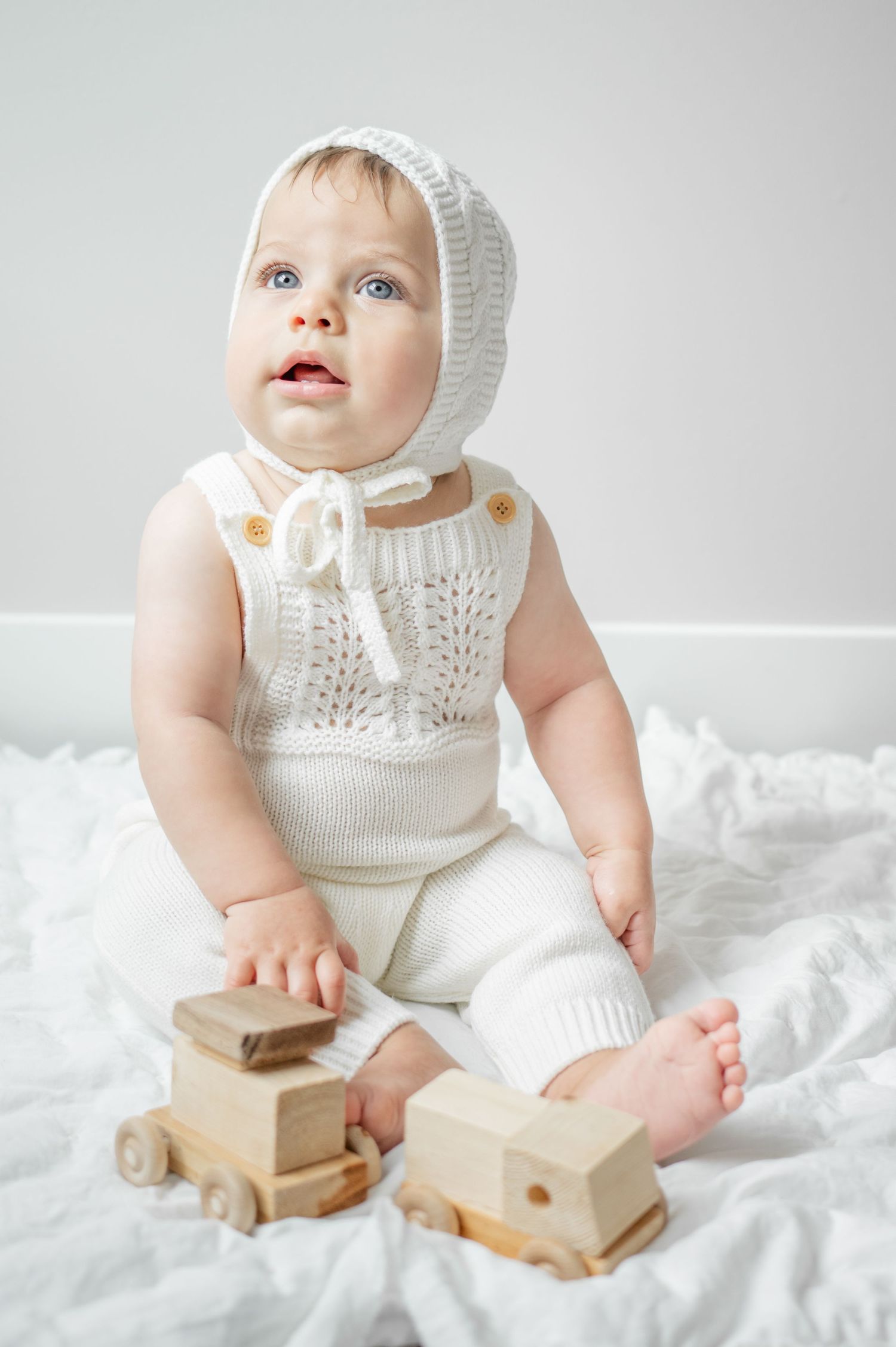 little boy in white knit outfit and bonnet looks up towards mom and dad while posing for images with little wooden cars