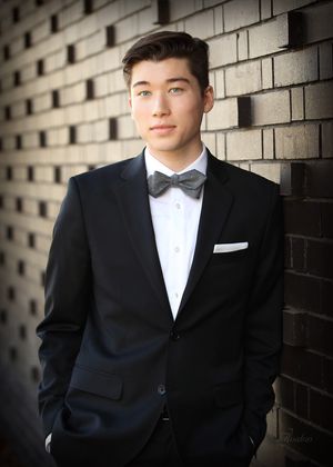 Formal portrait of a person in black tuxedo and bowtie against brick wall.
