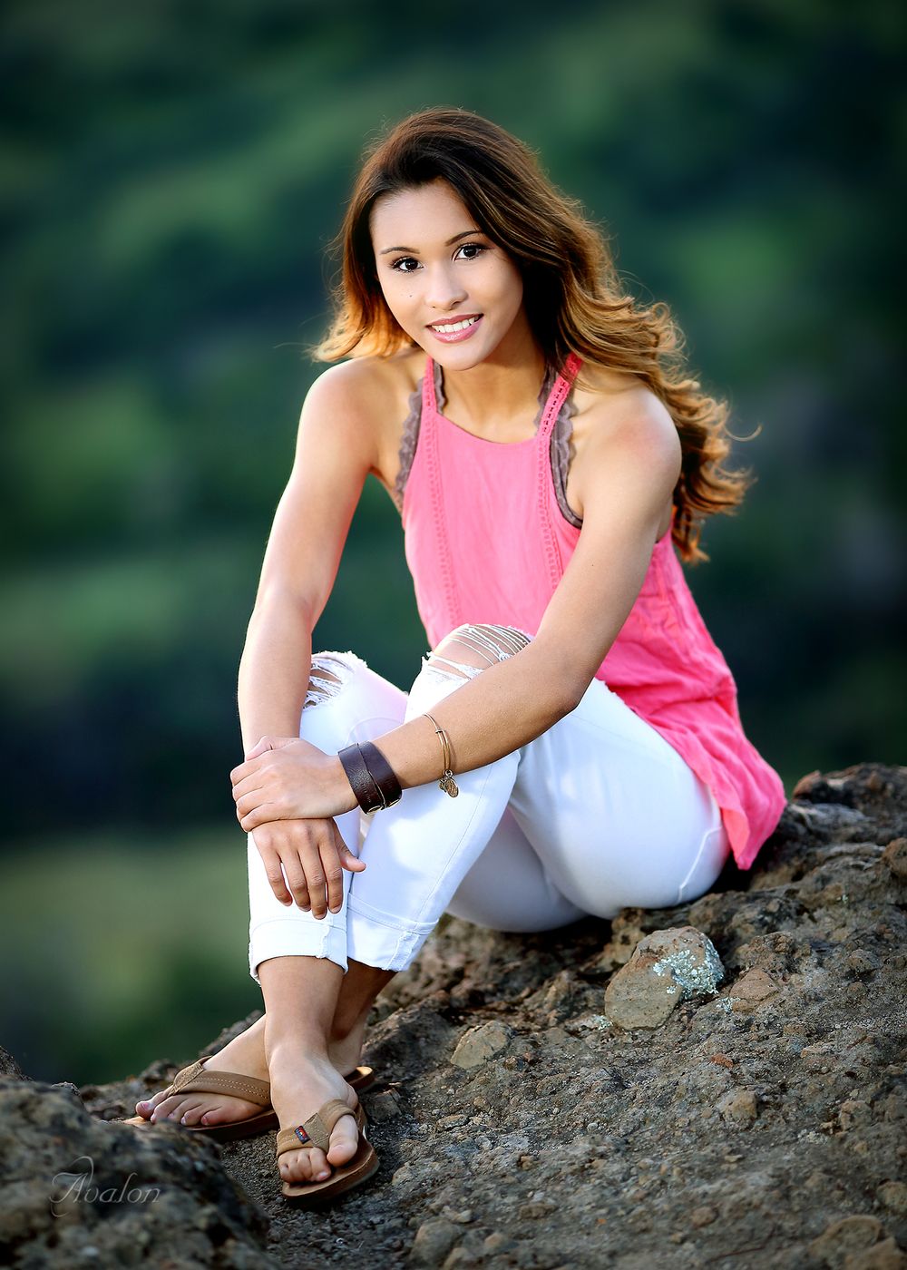 Outdoor portrait of someone in pink tank top and white pants sitting on rocks near water.
