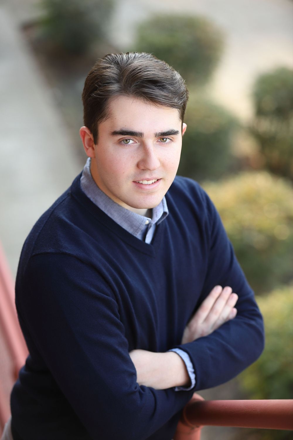 Young man in navy sweater and collared shirt poses with arms crossed against blurred background.