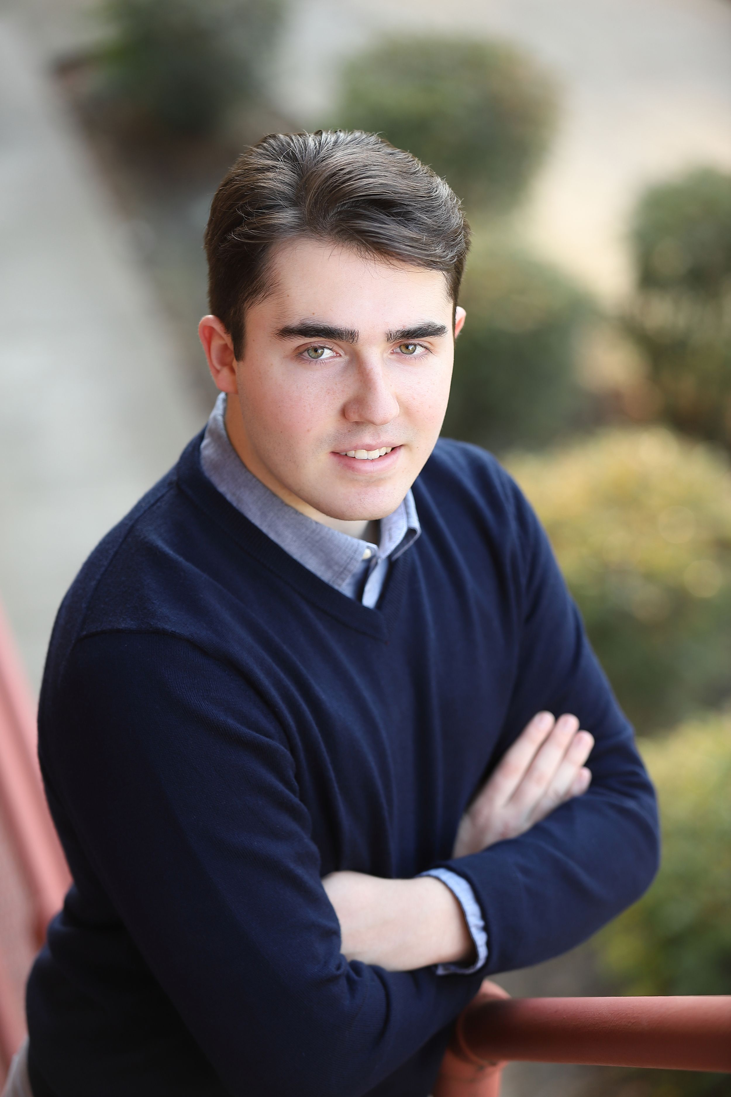 Young man in navy sweater and collared shirt poses with arms crossed against blurred background.