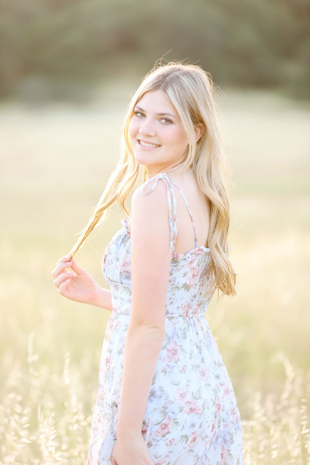 Woman in floral white sundress poses in sunlit field with flowing blonde hair.