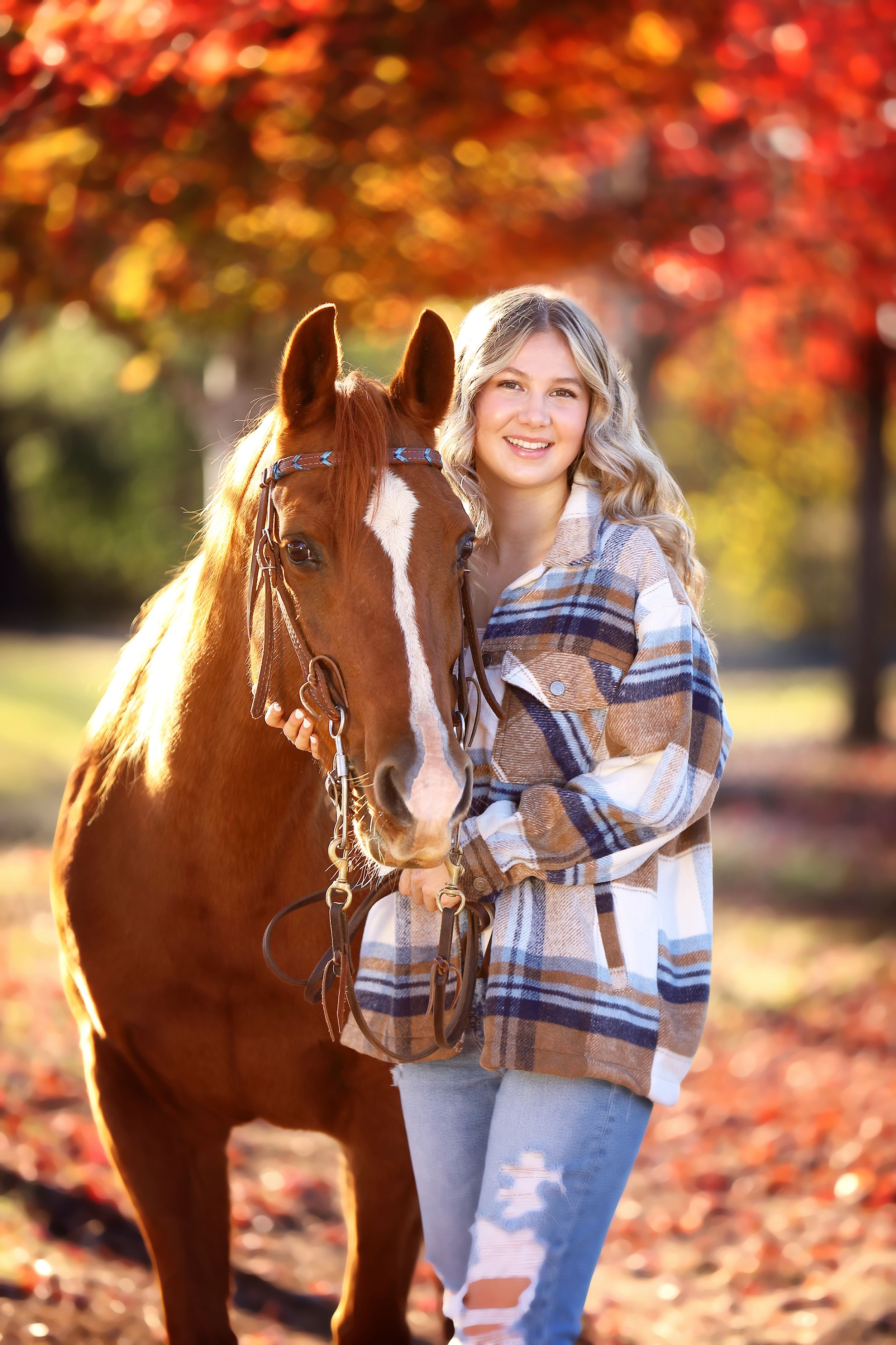 Person in plaid jacket stands with brown horse amid vibrant autumn foliage.