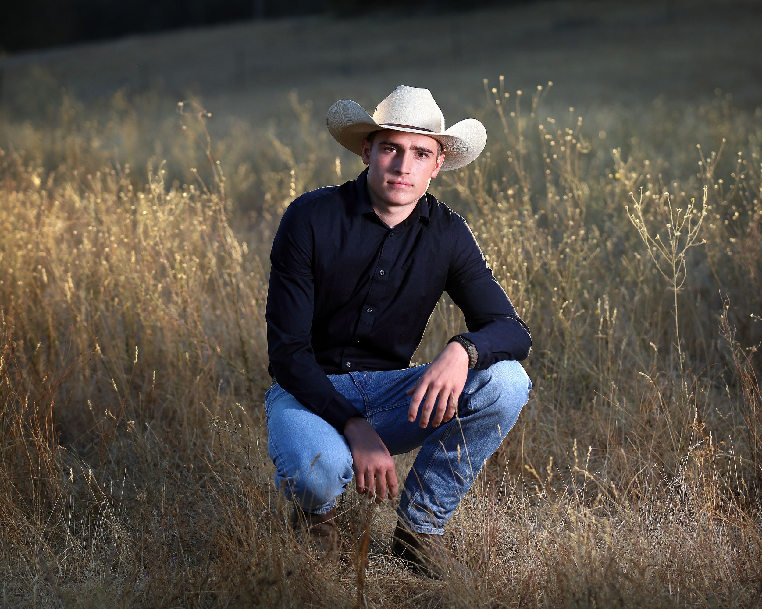 Portrait of person in cowboy hat and dark shirt crouching in tall grass at sunset.