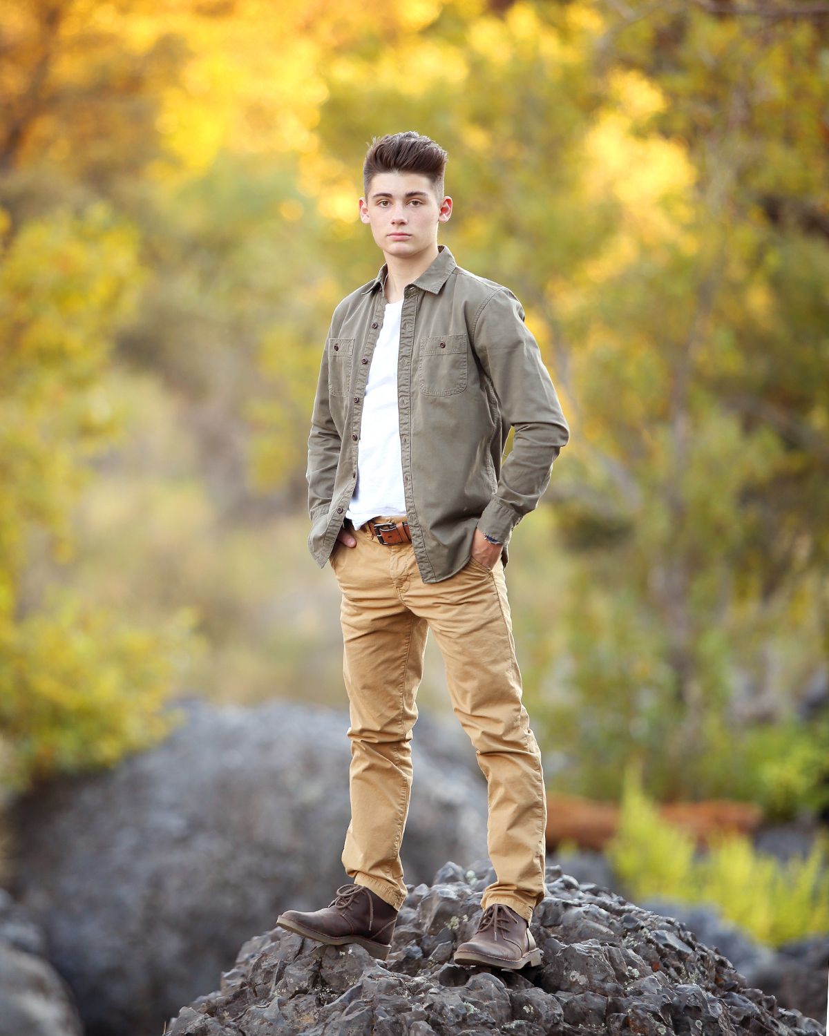 Young man in olive jacket stands confidently on rock with autumn colors in background.