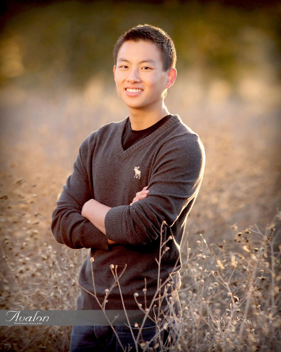 Young man in dark sweater poses with crossed arms in golden field at sunset.