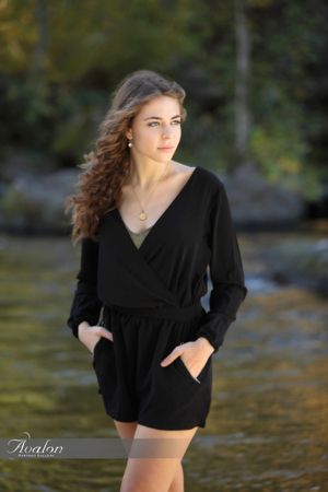Person in black romper poses by waterfront with long wavy brown hair.