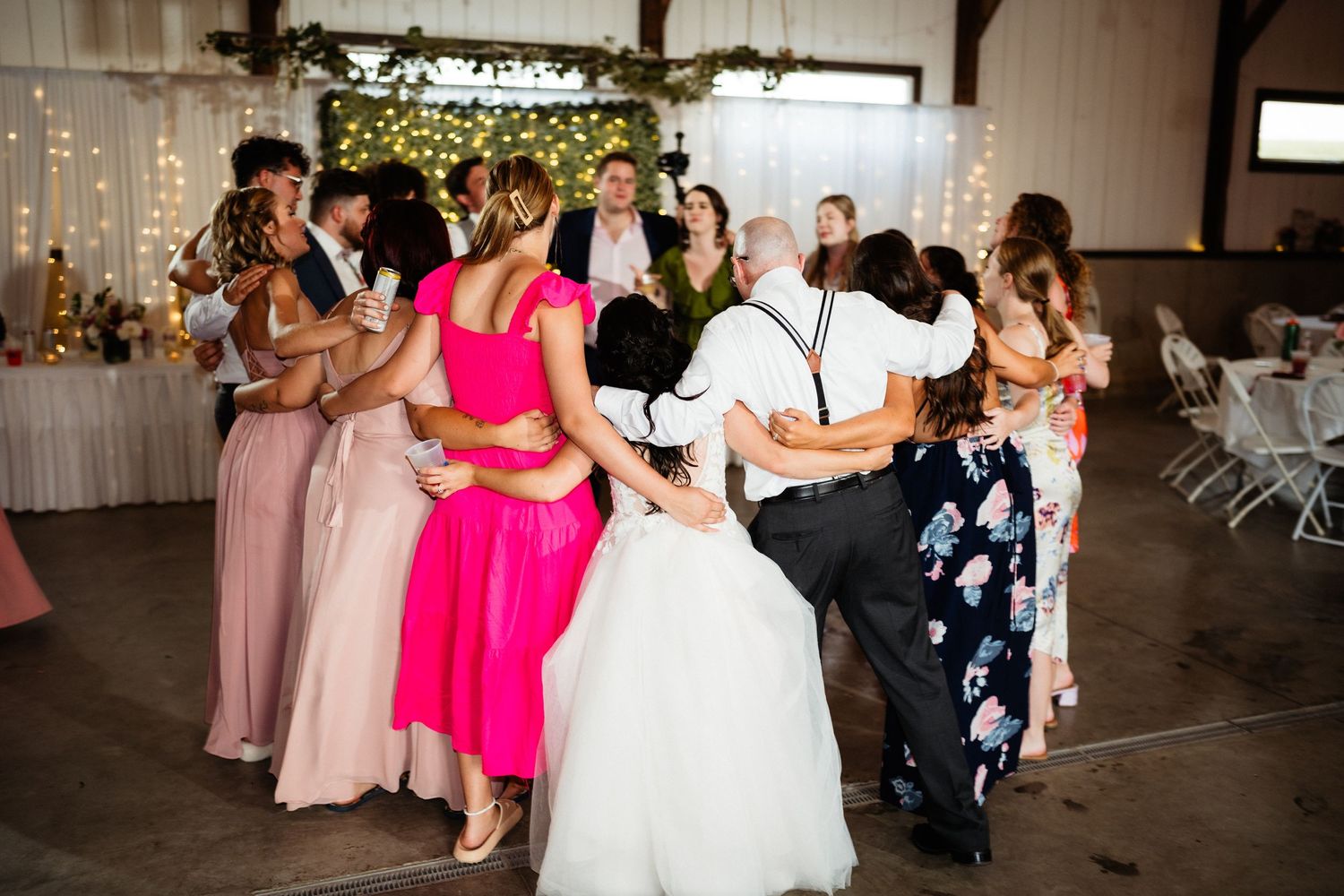 Wedding party dancing in a line at a reception venue with string lights and white drapes.