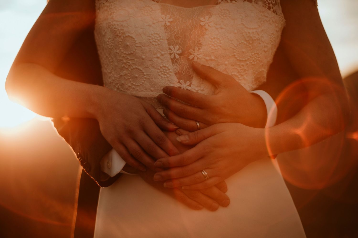 A close-up view of hands resting on a white wedding dress with warm sunlight creating a romantic glow.