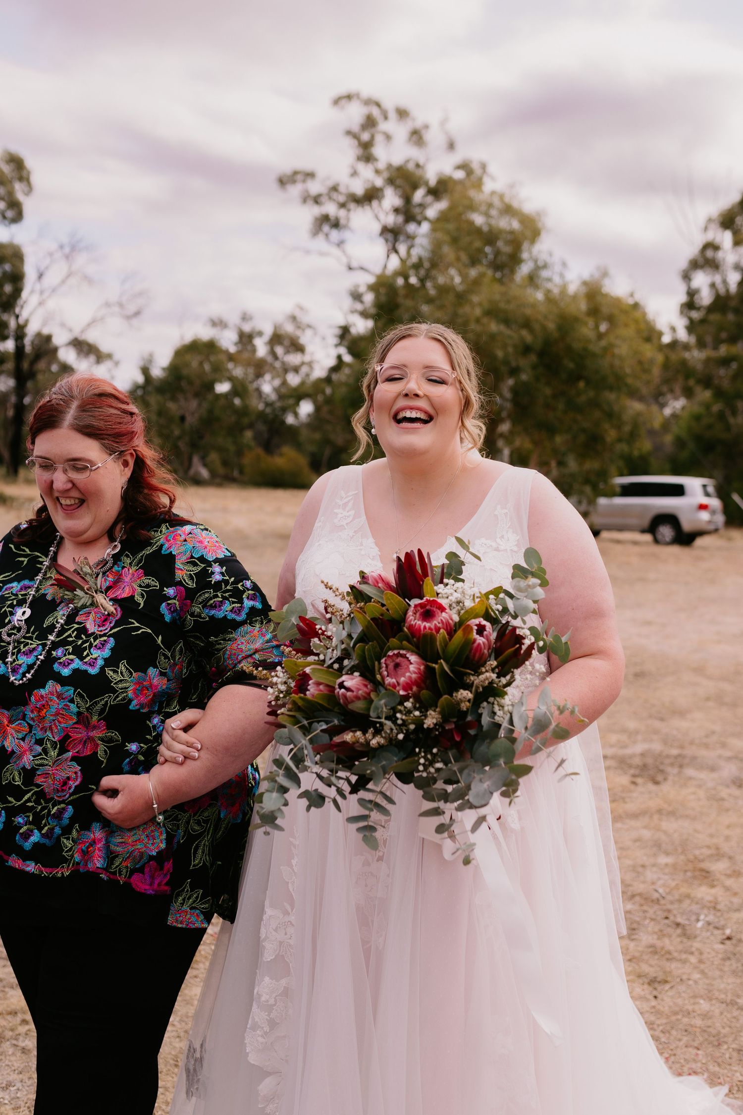 Joyful bride in white dress laughing while holding vibrant red and purple bouquet.