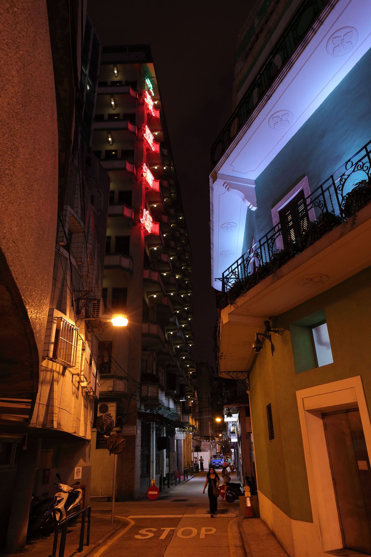 Narrow urban alley at night with tall buildings and red neon signs.
