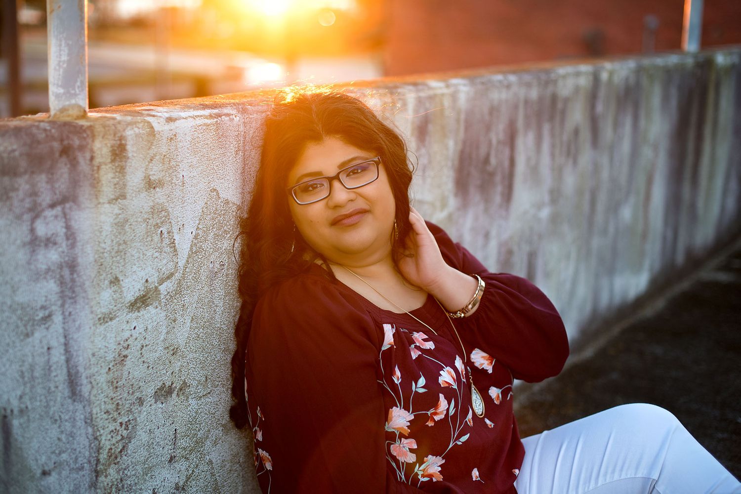 A young woman with glasses leans against a concrete wall during a golden sunset portrait session.