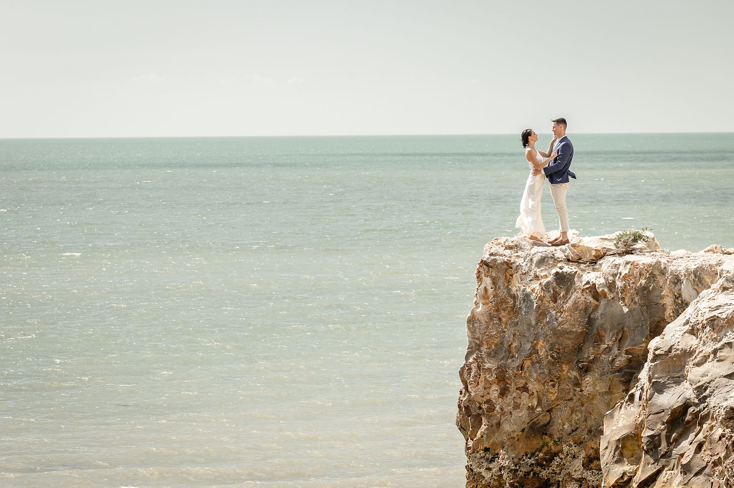 A couple stands at the edge of a dramatic seaside cliff in formal wedding attire.