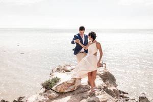 A couple in wedding attire stands together on coastal rocks overlooking the ocean.