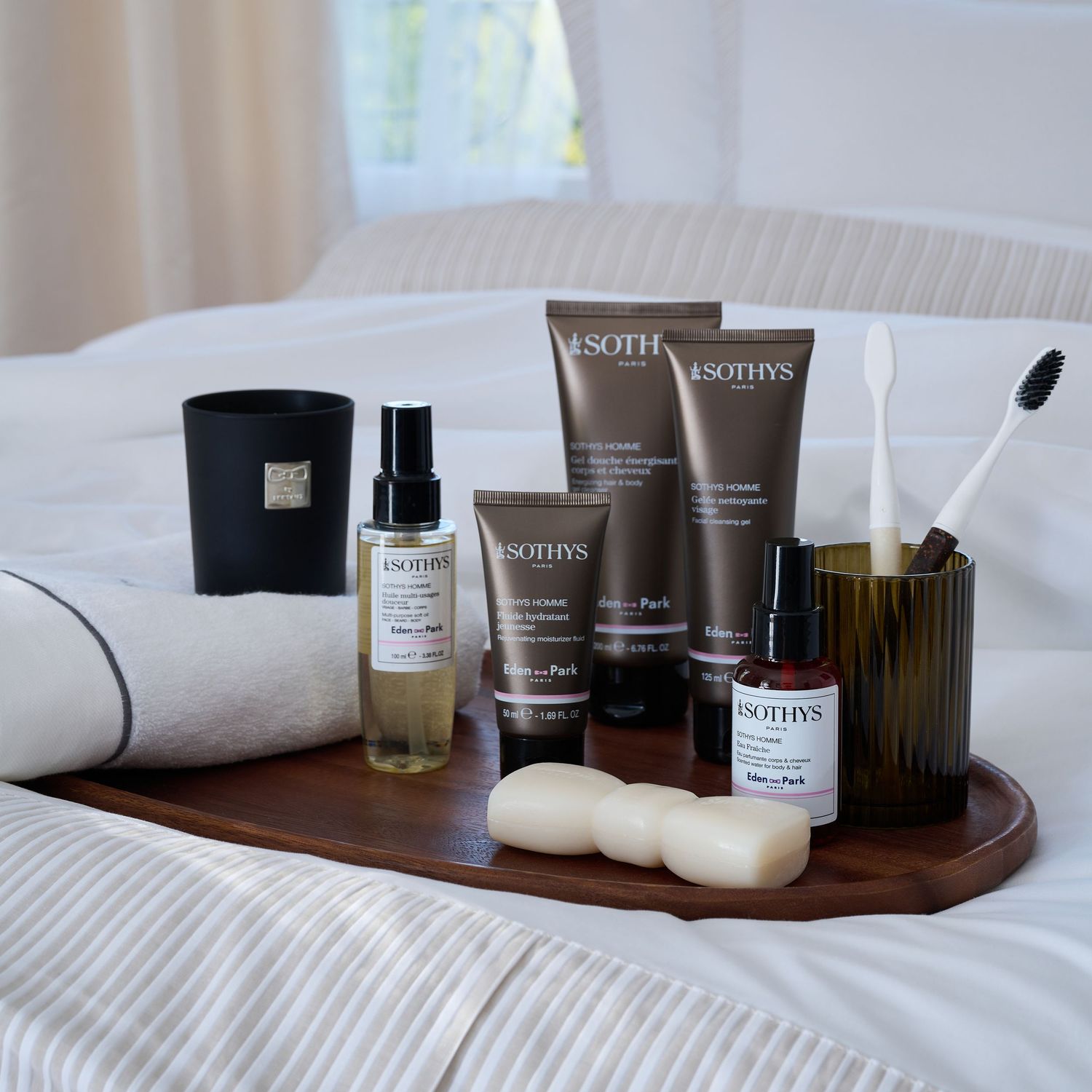 Collection of luxury skincare products arranged on wooden tray on white bedding.