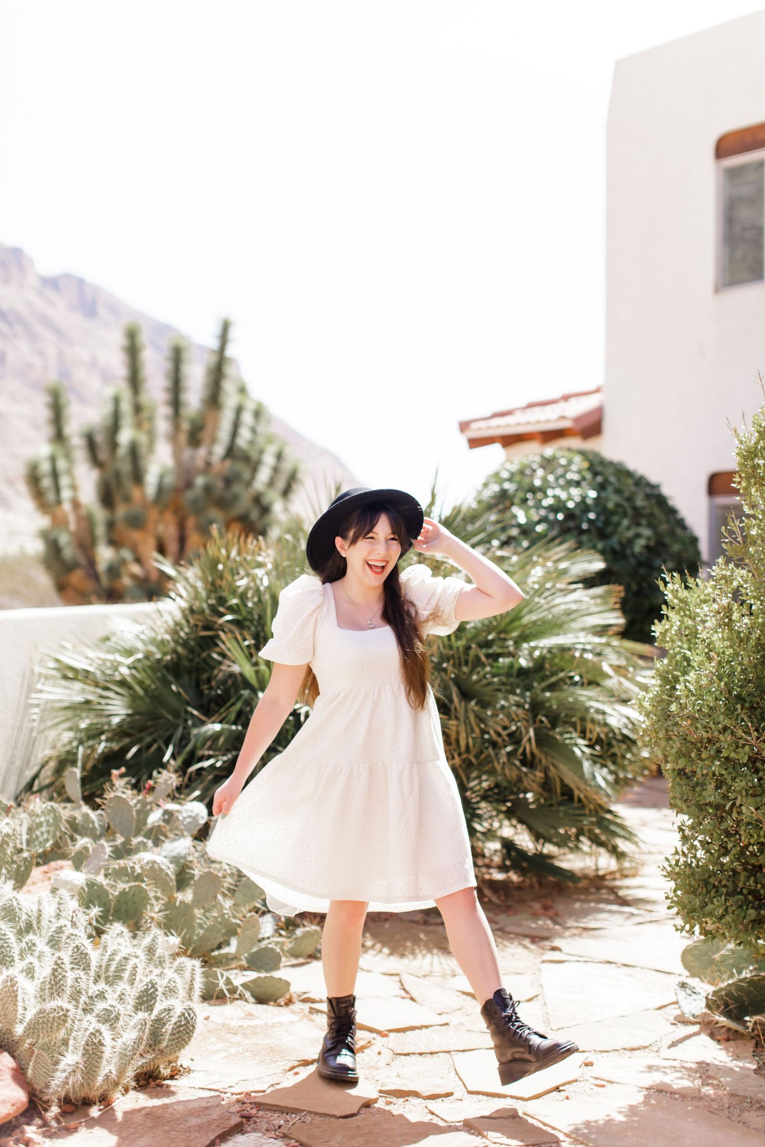 Danielle Debley Battistella in a white dress poses playfully in a desert garden surrounded by cacti and desert plants in Utah.