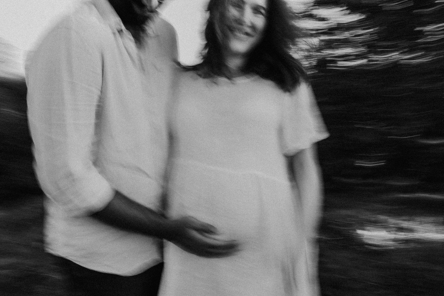 Black and white blurred artistic photo of a smiling pregnant couple embracing outdoors in white clothing.