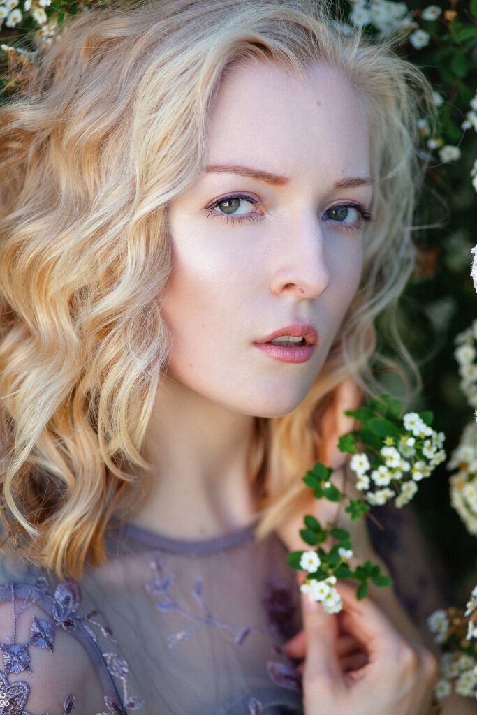 Portrait with wavy blonde hair and purple lace dress near white spring blossoms against natural foliage background.