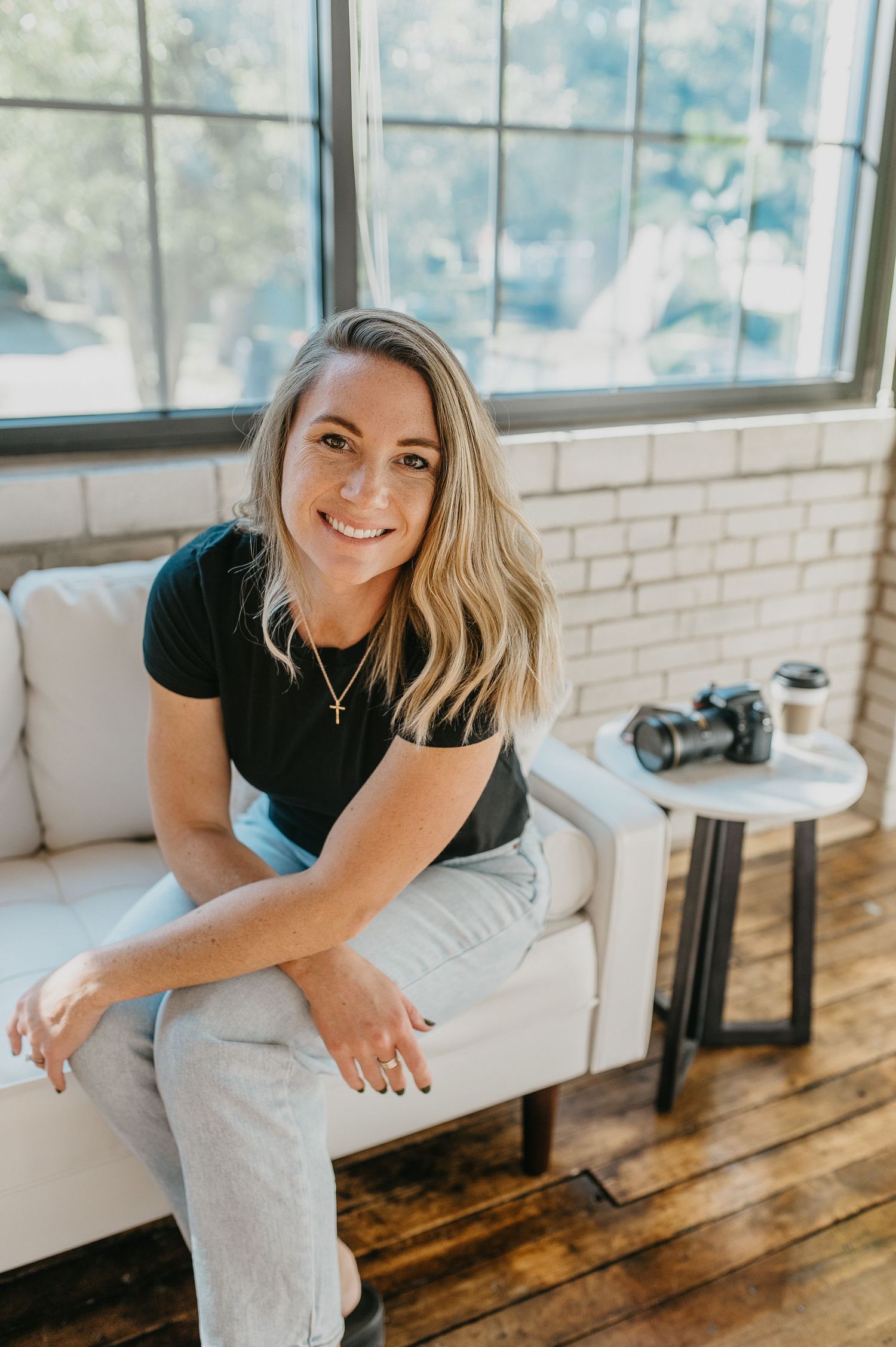 Casual portrait of woman sitting on white couch near window with camera on side table.