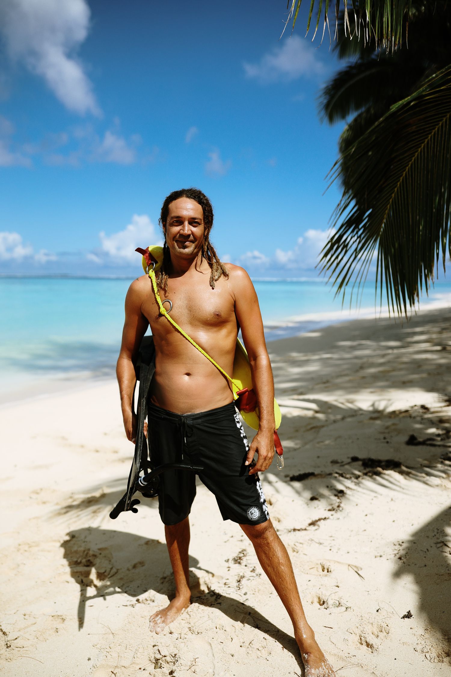 Beach goer poses with snorkeling gear on pristine white sand beach with palm fronds overhead.