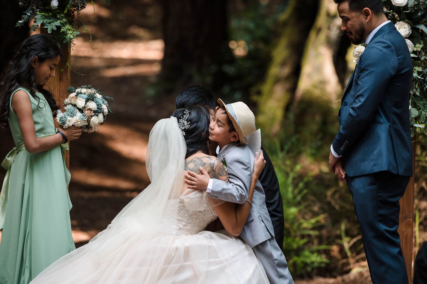 A tender moment shared during a woodland wedding ceremony with bridesmaids in sage green dresses.