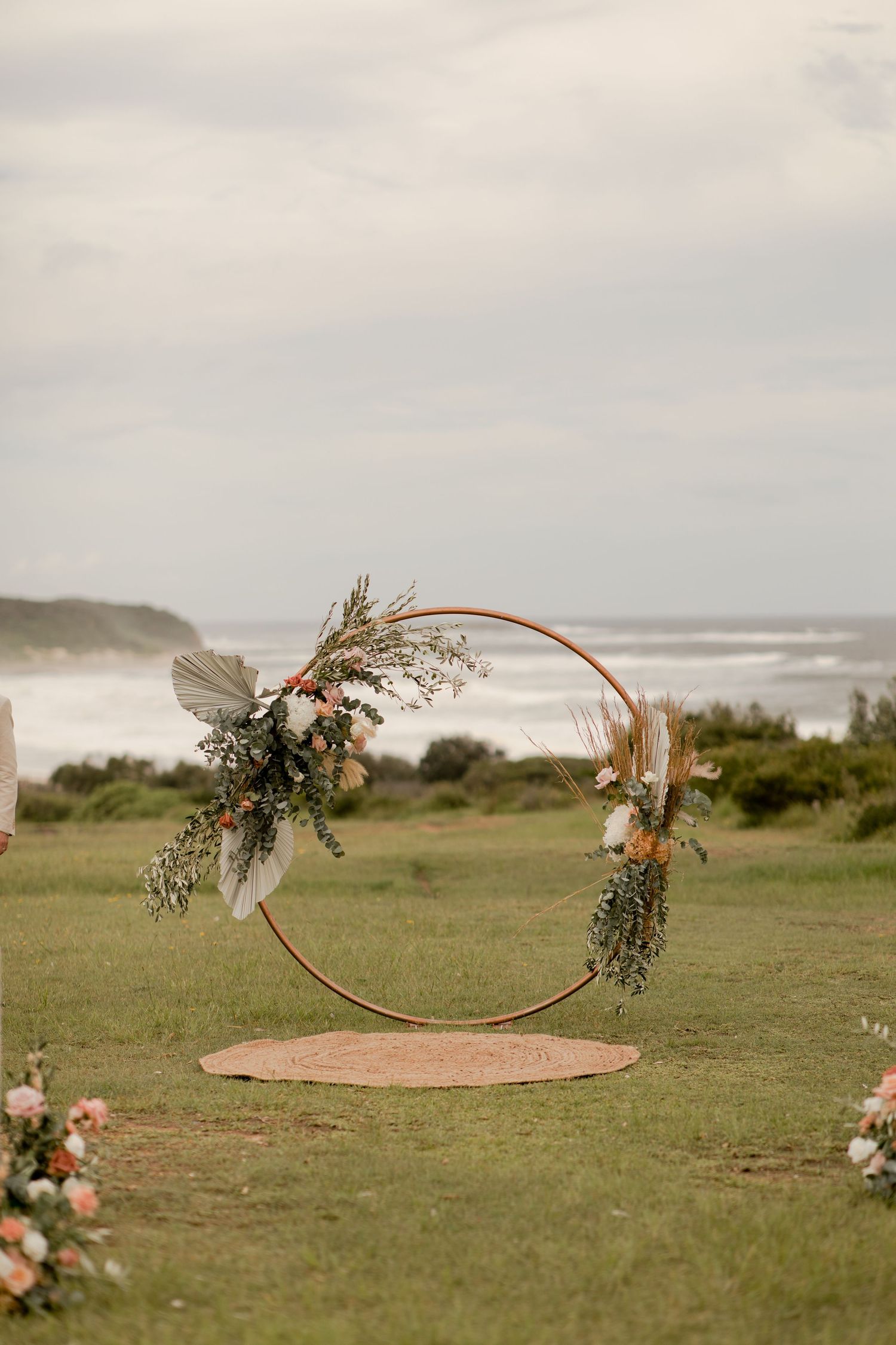 Circular wedding arch decorated with flowers and greenery overlooking a coastal landscape.