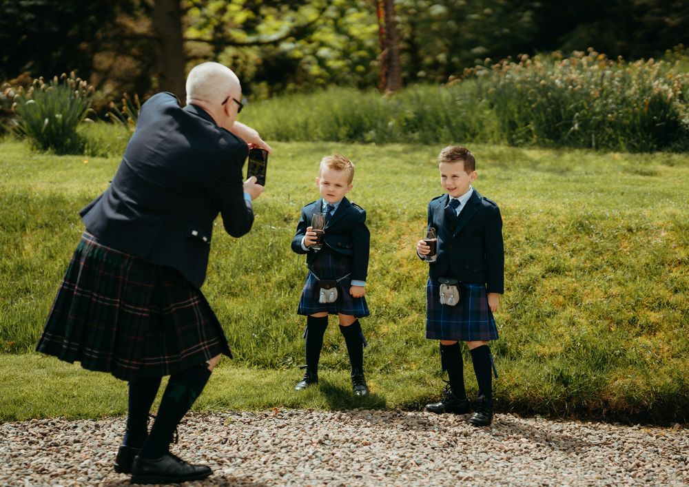 A photographer in a kilt captures young children posing on a grassy path surrounded by nature.