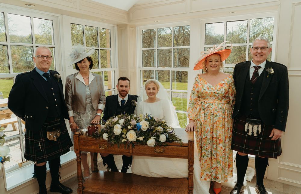 A formal group photo with traditional Scottish kilts and wedding attire in a bright indoor setting.