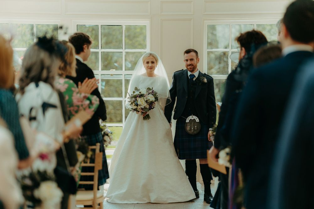 Wedding ceremony in a bright sunlit room with guests watching the couple at the altar.
