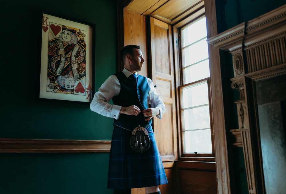 A person in traditional Scottish attire adjusts their outfit near a window in a dark green room.