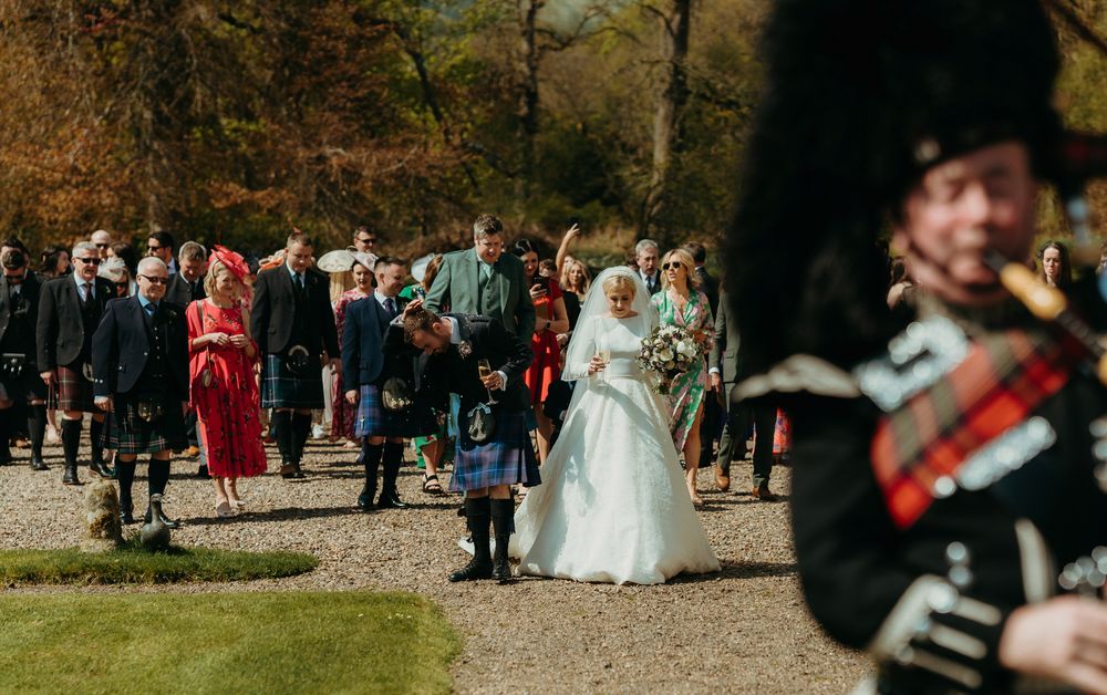 Wedding guests gather outdoors with bagpiper and traditional Scottish attire at a countryside venue.