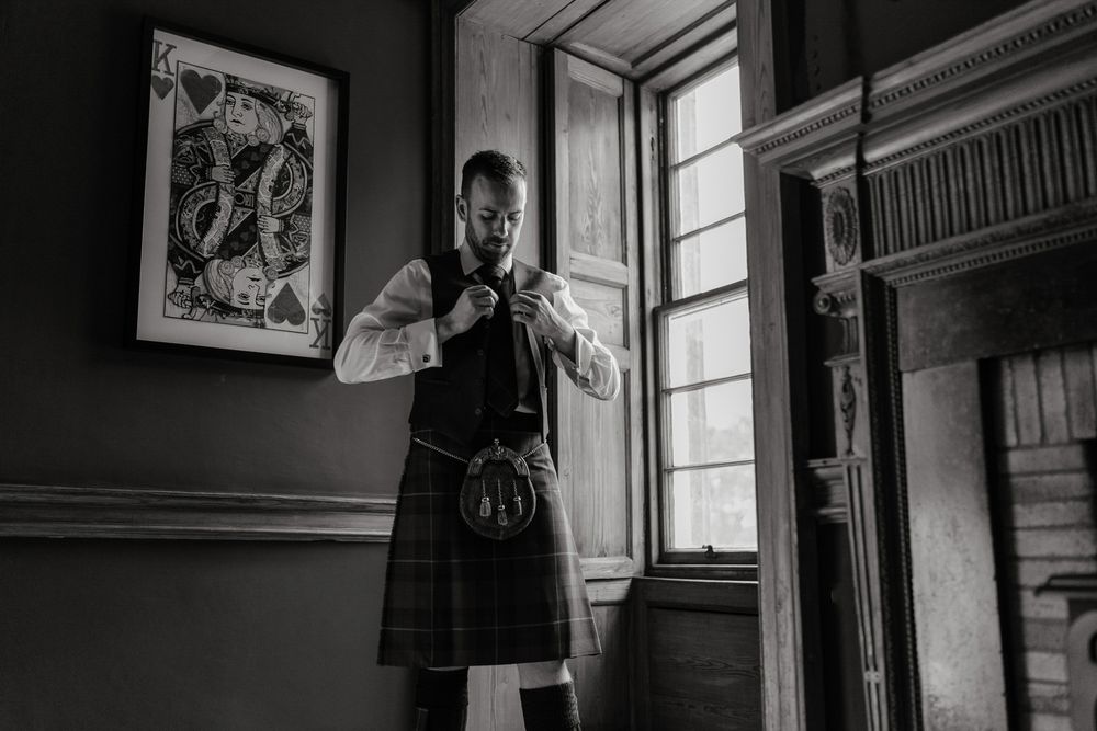 A silhouetted figure in traditional Scottish dress stands by a window in a dark room.