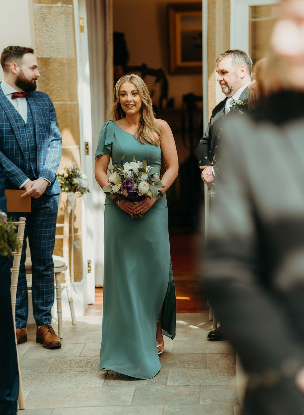 A bridesmaid in a sage green dress holds a floral bouquet while standing in an elegant hallway.