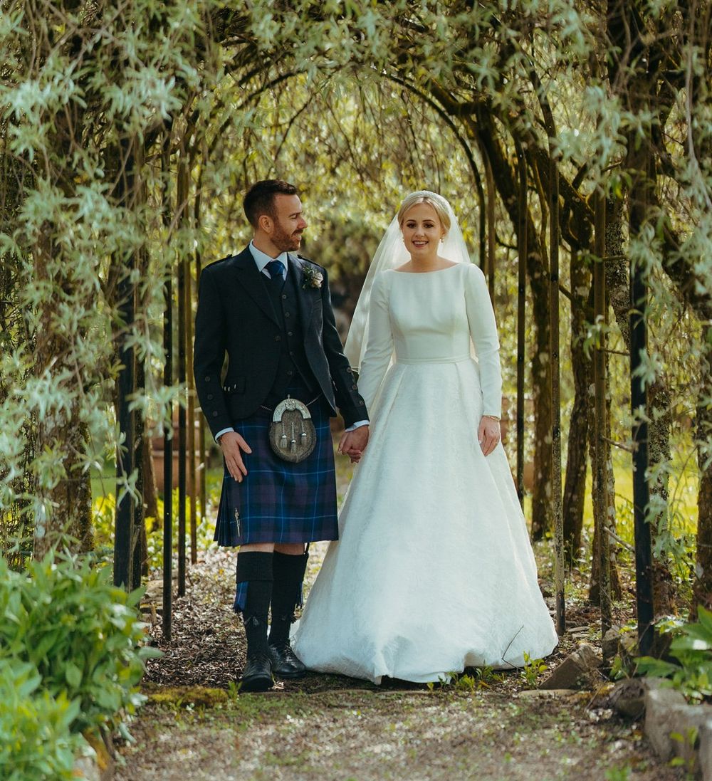 Newlyweds stand under a garden archway, with the groom in traditional Scottish kilt and bride in elegant white dress.