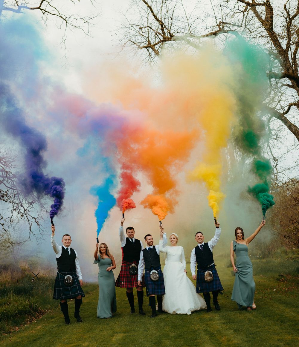 Wedding party holding colorful smoke bombs creates vibrant rainbow effect against bare winter trees.
