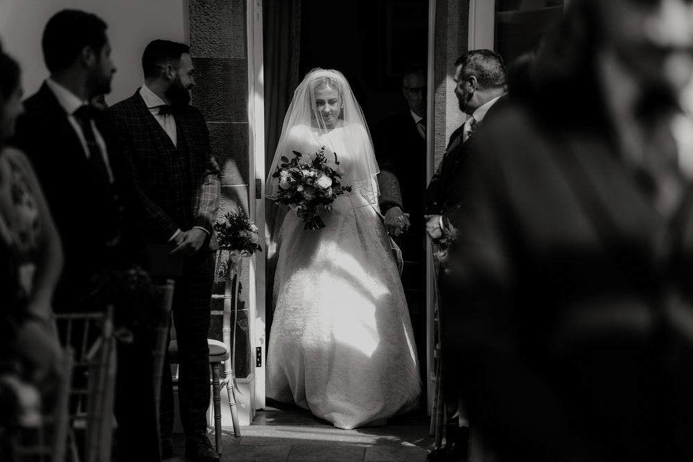 Black and white photo captures bride entering ceremony space through doorway.