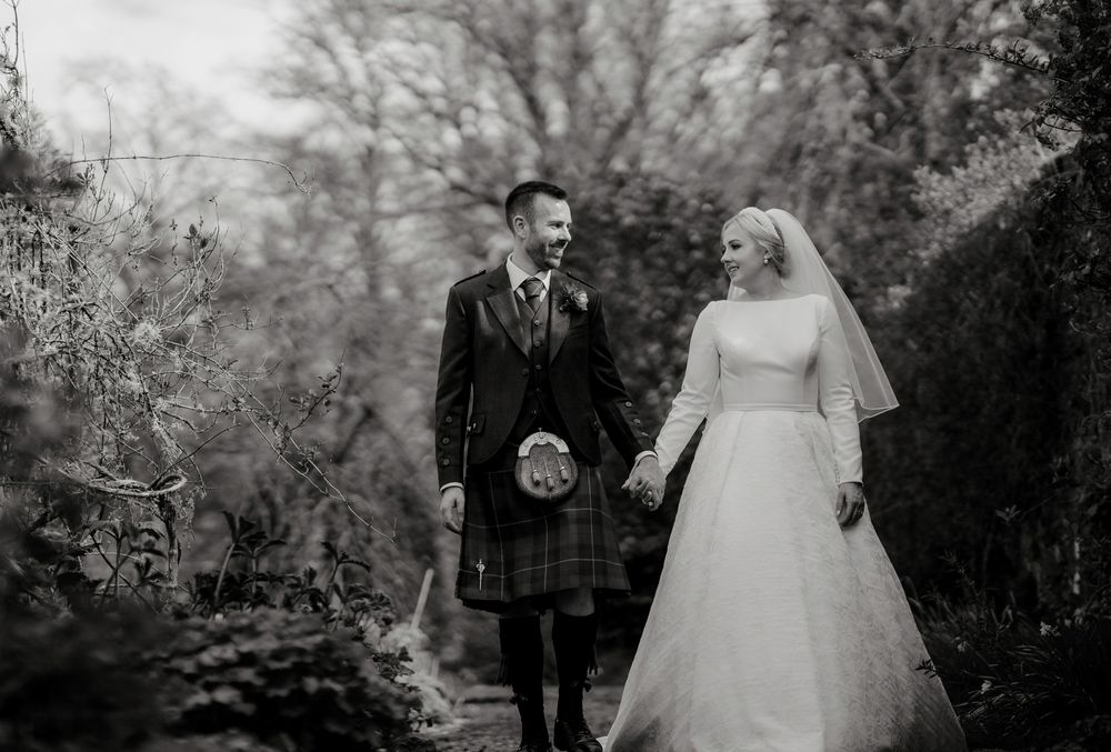 Black and white wedding portrait of couple walking through garden path in Scottish wedding attire.