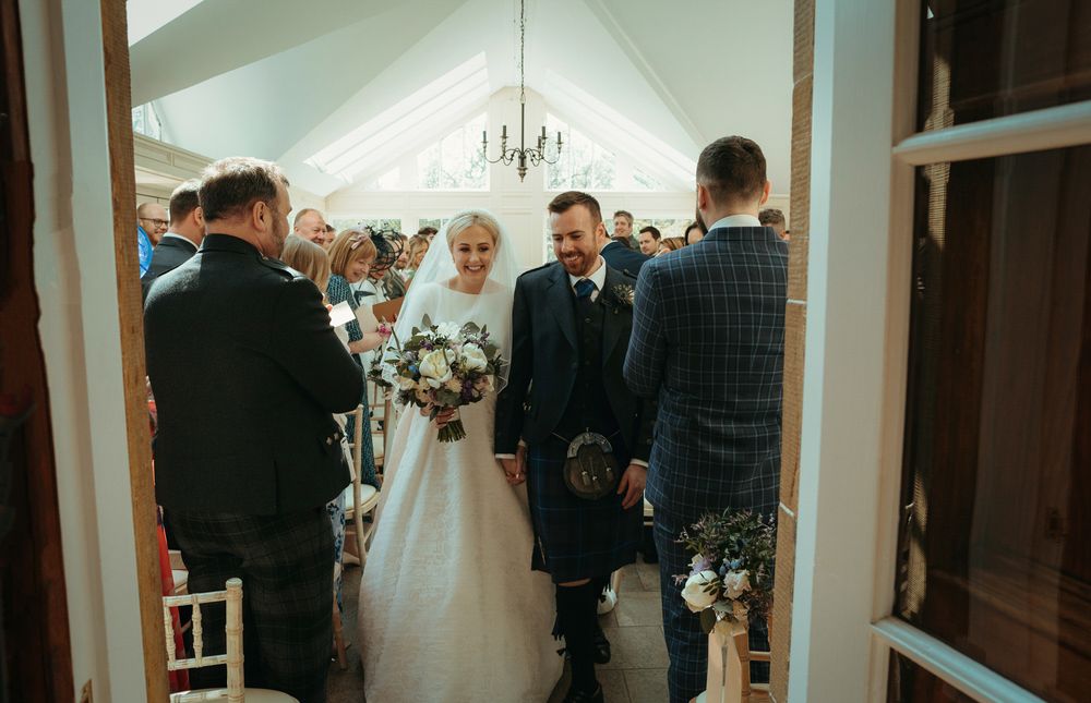 Wedding ceremony in progress viewed through doorway of bright white venue.